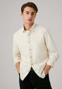 Crèmekleurig button-up shirt van katoen, met een puntkraag, houten knopen en opgerolde mouwen. Draagt over zwarte broek.