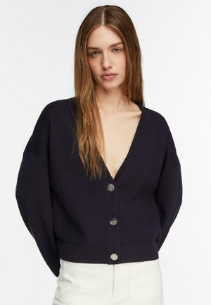 Femme aux longs cheveux bruns portant un cardigan noir à boutons avec un profond décolleté en V et un pantalon blanc, sur un fond blanc uni.