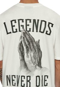 Fehér pamut póló, amelyen imádkozó kezek grafikája és a hátán található "A LEGENDÁK SOHA NEM HALNAK MEG" felirat vastag fekete betűkkel.