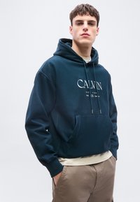 Νέος άνδρας που φοράει ένα σκουρομπλε φούτερ Calvin με κορδόνια και μπεζ παντελόνι, στέκεται με τα χέρια στις τσέπες μπροστά σε απλό φόντο.