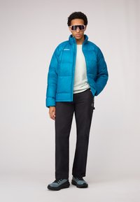 Veste bouffante bleu-vert avec un col montant, portée sur un haut vert clair. Pantalons noirs et chaussures de randonnée gris-bleu. Grandes lunettes de soleil blanches.