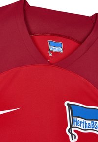 Czerwona koszulka sportowa z dekoltem w serek, wykonana z gładkiego materiału. Posiada białe logo Nike oraz niebieski herb Herthy BSC z grafiką flagi.