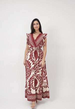 Mujer de pie vistiendo un vestido largo sin mangas de color crema con patrones de hojas y geométricos en rojo oscuro, posando frente a un fondo liso.