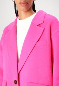 Ljust rosa ullblandad kappa med en notch-krage, en knappstängning och framfickor, bärs över en vit ribbad tröja.