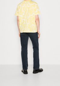 En person klädd i en mönstrad t-shirt i gult och vitt, mörkblå jeans och svarta läderskor, står mot en enfärgad vit bakgrund.