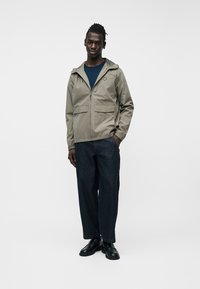 Άνδρας που στέκεται φορώντας μπεζ μπουφάν με κουκούλα, μπλε πουκάμισο, σκούρο τζιν με φαρδιά μπατζάκια και κόκκινη ραφή, και μαύρα παπούτσια, μπροστά σε λευκό φόντο.