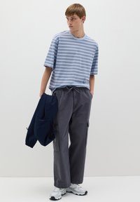 Maglietta a righe blu chiaro, fit morbido, abbinata a pantaloni cargo grigi scuri con tasche laterali, giacca blu navy in mano. Sneakers bianche completano il look.