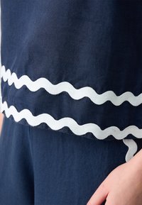 Marineblaues Textiloberteil mit weißem wellenförmigem Saum. Der Stoff wirkt weich und hat eine glatte Textur. Eine Hand ist am Rand sichtbar.