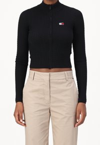 Top corto de punto acanalado negro con cierre y cuello alto y logo en contraste. Combinado con pantalones beige de tiro alto.