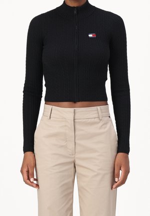 Haut zippé noir en tricot côtelé, court avec col montant et logo contrastant. Associé à un pantalon beige taille haute.