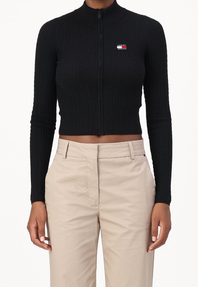 Top corto de punto acanalado negro con cierre y cuello alto y logo en contraste. Combinado con pantalones beige de tiro alto.