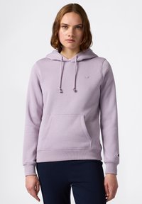 Giovane donna che indossa una felpa con cappuccio di un leggero colore viola con tasca frontale e pantaloni blu navy, in piedi contro uno sfondo bianco semplice.