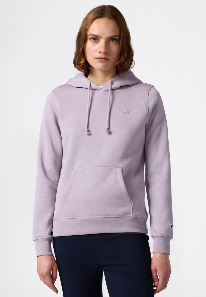 Giovane donna che indossa una felpa con cappuccio di un leggero colore viola con tasca frontale e pantaloni blu navy, in piedi contro uno sfondo bianco semplice.