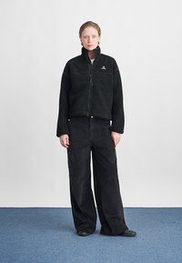 Črna zip-up flis jakna z visokim ovratnikom, strukturirano blago in majhnim logotipom. Usklajena s širokimi črnimi kord pantolonami in ravnimi čevlji.