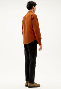 Braunes Cordhemd, taillierte Passform, Button-Down-Kragen, lange Ärmel, schwarze Hose, braune Wildlederschuhe, einfarbiger Hintergrund.