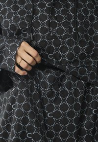 Zwarte jasje met een geometrisch patroon in grijs. Kenmerken zijn een rits, knopen en elastische manchetten. De textuur lijkt glad en glanzend.