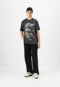 Young man wearing black cap, black graphic Marvel t-shirt, loose black jeans, and white sneakers standing against white background.