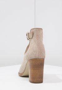 Beige mocka högklackad sko med rund tå, vadderad rem och silverspänne. Har en texturerad träklack och slät sula.