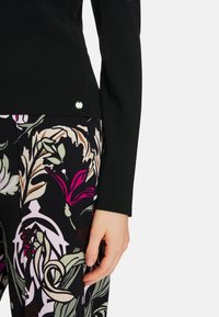 Schwarzer Pullover mit langen Ärmeln und einem kleinen Logodetail am Saum, kombiniert mit eng sitzenden Hosen mit einem mehrfarbigen Blumenmuster auf dunklem Hintergrund.