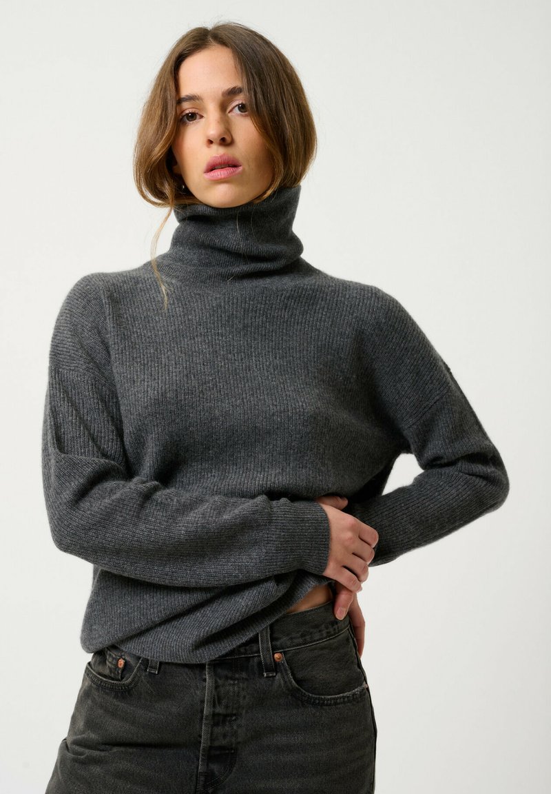 Grijze geribbelde coltrui met lange mouwen, gemaakt van zacht materiaal, gecombineerd met donkere jeans. De pasvorm is ontspannen en comfortabel.
