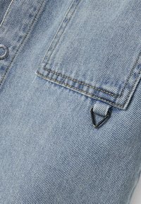 Svetlo modra denim tkanina z teksturirano površino, ki ima žep na prsih in majhen trikotni črn kovinski detajl.