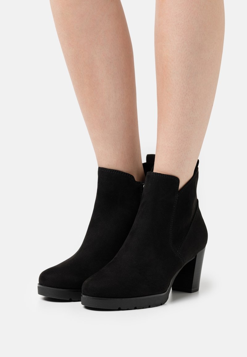 Paire de bottines en suède noir à talons blocs portées sur des jambes nues sur un fond clair uni.