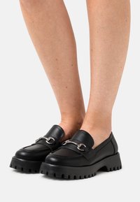 Mocasines de cuero negro con suela gruesa y estriada, que presentan un acento de cadena plateada en la parte superior y una textura suave.