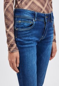 Plonėjimo įdubimo mėlynos spalvos džinsai su penkių kišenių dizainu, matomomis siūlėmis ir tekstūruotu paviršiumi. Dėvimi su margintu ilgomis rankovėmis viršutiniu drabužiu.