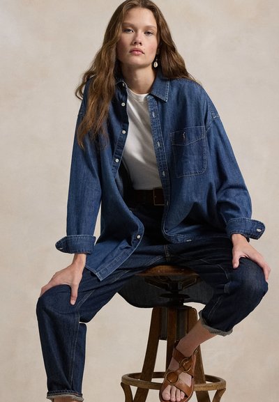 Junge Frau sitzt auf einem Holzschemel, trägt ein übergroßes Jeanshemd, ein weißes T-Shirt, Jeans und braune Sandalen, neutraler Hintergrund.