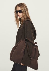 Frau in einem dunklen, oversized Pullover und Sonnenbrille, die eine große braune Wildlederhandtasche über die Schulter trägt.