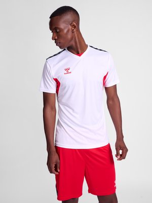 Mann trägt ein weißes Sporttrikot mit roten Akzenten und roten Shorts, schaut nach links vor einem einfarbigen Hintergrund.