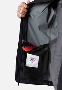 Das Innere einer schwarzen Jacke zeigt einen grauen Futterstoff, eine Netztasche mit einer Sonnenbrille und ein beschriftetes Patch mit einem rot-blauen Logo.