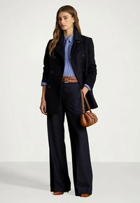 Nő fekete, dupla soros kabátot visel egy kék csíkos ing felett, fekete csíkos, bő szárú nadrággal, barna övvel, cipővel és egy barna táskával a kezében.