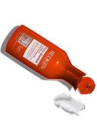 Redken Frizz Dismiss, bottiglia di plastica arancione, con pompa, posata di lato. Prodotto cremoso bianco spremuto vicino, texture liscia.