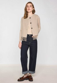 Gilet lavorato a maglia beige con orlo frangiato, tre bottoni, indossato sopra una maglietta chiara e jeans scuri, caratterizzato da maniche risvoltabili e mocassini marroni.