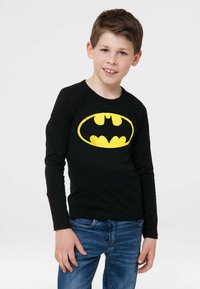 Schwarzes Langarmshirt mit großem gelben Batman-Logo. Kombiniert mit blauen Jeans, vor einem weißen Hintergrund.