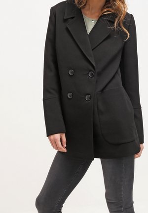 Frau trägt einen schwarzen, doppelt geknöpften Blazer mit großen Vordertaschen und dunkelgraue, figurbetonte Jeans vor einfarbigem Hintergrund.