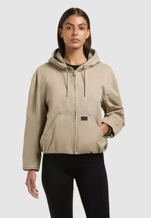 Fiatal nő bézs színű, elején zsebekkel ellátott cipzáras kapucnis dzsekit és fekete leggingset visel, egyszerű világos háttér előtt áll.