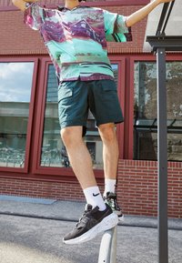 Person balancing on a metal rail outdoors wearing black Nike sneakers, white socks, dark shorts, and a colorful patterned shirt.
