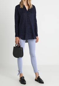 Marineblauwe blouse met lange mouwen in textuurstof, lichtblauwe aansluitende broek met zijbanden, zwarte flatschoenen en een kleine zwarte handtas.