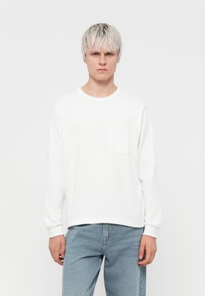 White long-sleeve t-shirt made of cotton, featuring a round neckline and a small pocket on the left chest. Paired with light blue jeans.