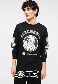 Μαύρο T-shirt με μακριά μανίκια, με κεντρικό γραφικό που απεικονίζει μια γυναίκα και παγωτό, με λευκό κείμενο και σχέδια στα μανίκια. Απαλή υφή υφάσματος.