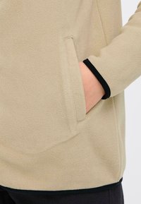 Beige fleecejack met een zijzak en zwarte rand bij de manchetten, met een zachte textuur en een ontspannen pasvorm ontwerp.