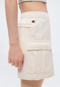 Φούστα cargo σε κρεμ απόχρωση με εμπρόσθια τσέπη με καπάκι και μικρό κεντητό λογότυπο κοντά στη ελαστική μέση σε χαμηλό τμήμα κορμού ατόμου.