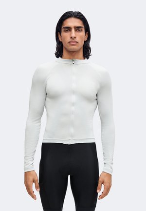 Homme aux cheveux foncés mi-longs, portant un haut zippé ajusté à manches longues gris clair et un cuissard noir, debout devant un fond uni.