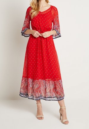 Nainen, jolla on päällään punainen midimekko paisley-kuviollisella reunuksella, läpikuultavilla kolmanneksen hihoilla, vyötäröltä nyörityt yksityiskohdat ja beiget korkokengät.