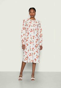 Vestido floral com base branca, featuring flores vermelhas e azuis. Com um colarinho de babados e mangas longas, até ao joelho. Usado com sandálias.