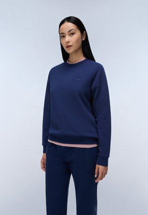 Kvinde med langt mørkt hår iført en mørkeblå sweatshirt og bukser over en lyserød skjorte, stående mod en ensfarvet lys baggrund.