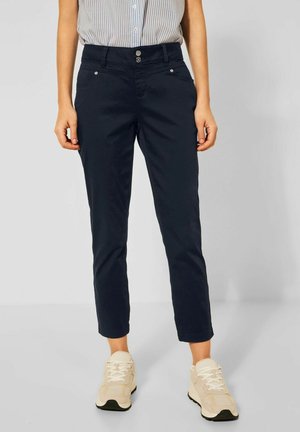 Femme portant un pantalon court bleu marine, des baskets beige et une chemise à rayures bleu clair rentrée, debout devant un fond uni.
