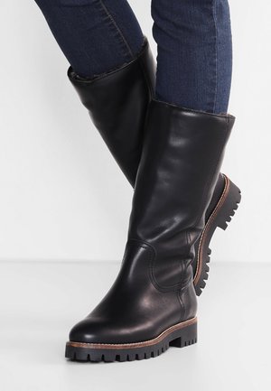 Person iført sorte læderstøvler til midtkalven med profilerede såler og brune kanter, kombineret med mørkeblå denimjeans.